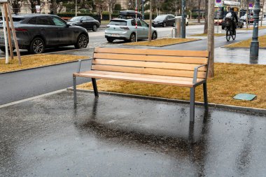 Viyana - 21 Şubat 2026: Temiz ahşap bir bank, araçlar ve yeşilliklerle çevrili yağmurdan sonra ıslak bir kaldırımda duruyor.