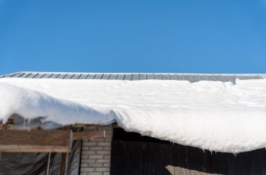 Kar, açık mavi gökyüzü altında güneş ışığını yansıtan metal bir çatının kenarını kaplıyor. Kırsal bir ortamda kış havası, mevsimlik temalar ve açık hava görüntüleri için mükemmel..