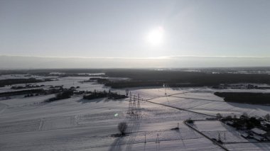 Açık bir gökyüzünün altında parlak bir güneşle karla kaplı bir manzaranın havadan görünüşü. Sahnede açık tarlalar, ağaç siluetleri ve huzurlu bir kış atmosferini çağrıştıran uzak tepeler yer alıyor..