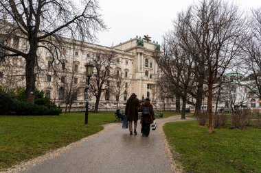 Viyana - 21 Şubat 2026: Arka planda çıplak ağaçlar ve tarihi mimariyle çevrili bir parkta yürüyen üç kişi. Atmosfer sakin..