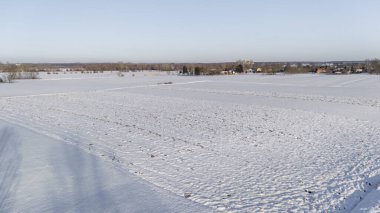 Açık mavi gökyüzünün altında, arka planda sakin kırsal güzelliği gösteren uzak ağaçlar ve evler ile kaplı, geniş tarlalarla kaplı kış manzarası..