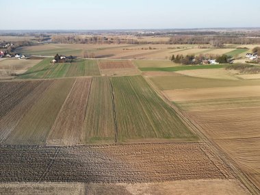 Açık gökyüzünün altında yeşil ve kahverengi ekin desenleri gösteren tarımsal alanların havadan görünüşü. Bu manzara tarım arazilerinin, tarım kültürlerinin, kırsal yaşamın önemini ön plana çıkarıyor..