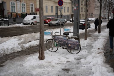 Viyana - 21 Şubat 2026: Üzerinde reklamlar olan park edilmiş bir bisiklet, karlı bir manzaranın ortasında kış kentsel yaşamını gözler önüne seren bir caddede karla kaplıdır.