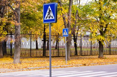 Sonbaharda bir parkta ağaçlarla kaplı bir caddenin yanında iki yaya geçidi tabelası duruyor. Sarı yapraklar zemini kaplıyor, resim gibi bir manzara yaratıyor..
