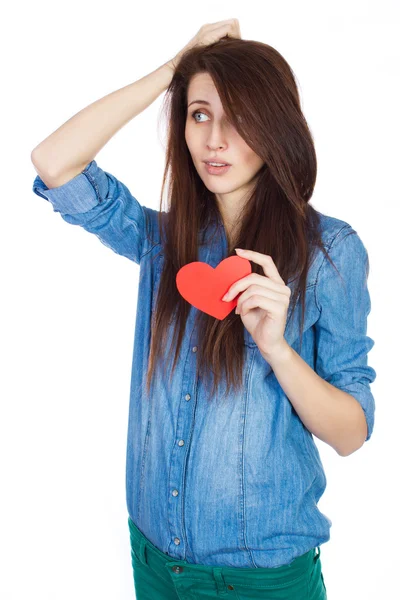 Smuk ung pige i denim blå skjorte stående på en hvid baggrund med et rødt papir hjerte i hænderne . - Stock-foto