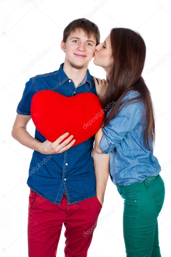 Eautiful young happy couple kissing behind a red heart, holding it in hands, isolated on a white background