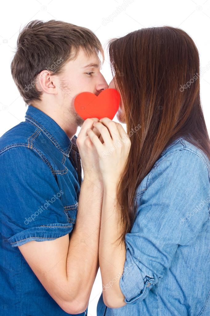 Beautiful young happy couple kissing behind a red heart, holding it in hands,  isolated on a white background