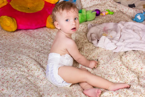 Schattige blonde babymeisje met mooie blauwe ogen zit op bed in luiers met speelgoed Stockfoto