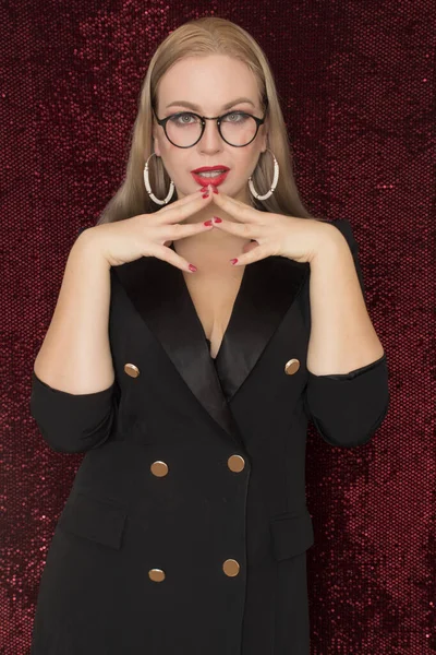 赤い光沢のある背景にポーズをとった黒いドレスジャケットの眼鏡の美しい若いブロンドの女性 彼女の頬にハート — ストック写真
