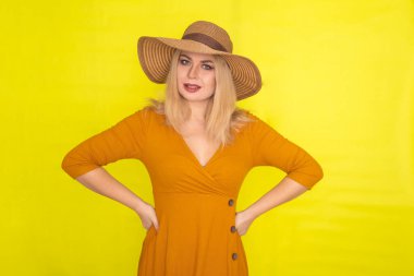 Sarı arka planda poz veren kahverengi şapkalı ve koyu sarı elbiseli güzel sarışın kadın. Moda yaz görünümü