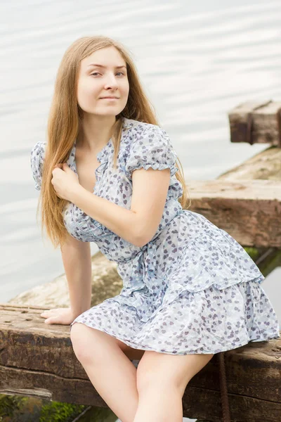 Portrait d'une belle jeune femme aux longs cheveux bruns dans une robe courte avec un motif floral. Fille est relaxant à l'étang sur des poutres en bois — Photo