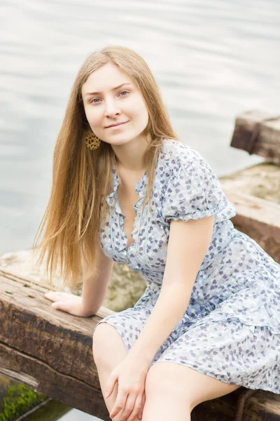 Retrato de una hermosa joven con el pelo largo y castaño en un vestido corto con un patrón floral. Chica es relajarse en el estanque en vigas de madera —  Fotos de Stock