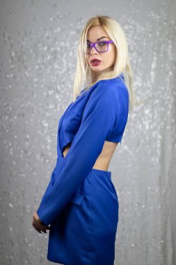 Stüdyoda, parlak gümüş arka planda mavi ceket ve mor gözlüklü sarışın kadın portresi. Moda ve gözlük konsepti.