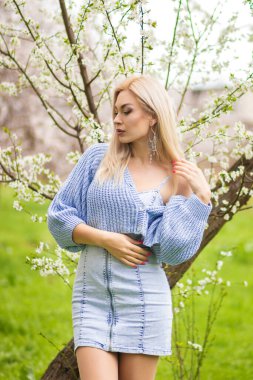 Açık havada beyaz çiçeklerin arasında sarışın bir kadın. Mavi örgü hırka ve kot elbiseyle bahar moda portresi..