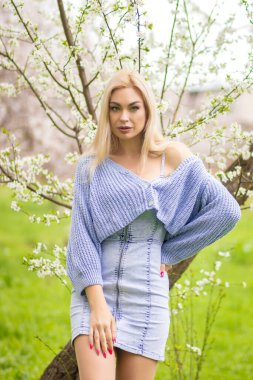 Çiçek açan ağacın yanında el ele tutuşan sarışın manken. Mavi örgü hırka ve kot elbise içinde bahar portresi, doğal ışık..