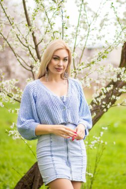 Bahar bahçesinde beyaz çiçeklerle gülümseyen sarışın kadın. Mavi hırka ve kot elbise içinde sıradan moda portresi, doğal gün ışığı..