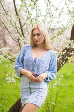 Sarışın kadın, ilkbaharda çiçek açan ağacın yanında güven içinde poz veriyor. Açık hava yaşam tarzı portresi, mavi örgü süveter, kot elbise, bokeh.