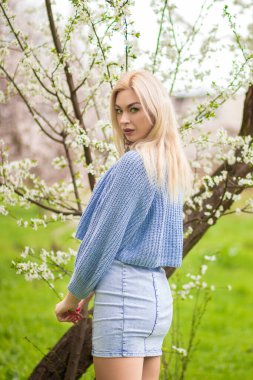 Arkaya bakan sarışın kadının çiçek açan ağacın arkasından bakışı. Mavi örgü hırka ve kot elbise içinde bahar moda portresi