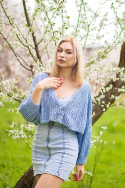 Zarif sarışın kadın çiçek açan ağacın yanında boyuna dokunuyor. Mavi örgü hırka ve kot elbise içinde bahar yaşam tarzı portresi, sığ alan derinliği.