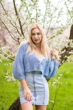 Sarışın kadın dalların yanında hafifçe gülümsüyor. Bahar güzeli ve mavi örgü hırka ve kot elbise içinde moda portresi.