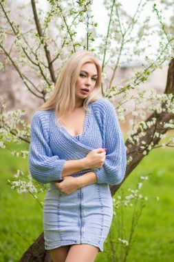 Dalgın sarışın kadın çiçek açan bir ağacın yanında kendine sarılıyor. Yeşil bir bahçede sakin bir bahar portresi.