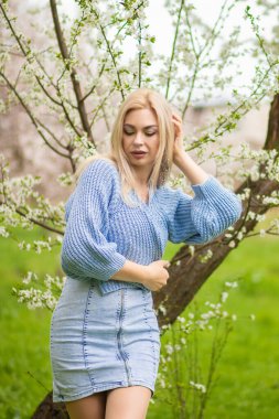 Genç sarışın kadın çiçek açan bir meyve bahçesinde saçına dokunuyor mavi kazak ve kot elbise giyiyor. Açık havada rüya gibi bahar moda portresi.