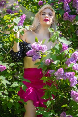 Çiçek açan leylak çalılarında poz veren mutlu sarışın kadın, açık havada mor çiçekler, yeşil yapraklar ve güneş ışığı olan bir portre..