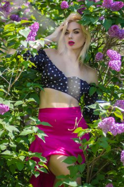 Sarışın kadın elinde akıllı bir telefonla güneşli bir bahçede leylak çiçeklerinin arasında poz veriyor. Sosyal medya, mesajlaşma ve bahar güzelliği konsepti.