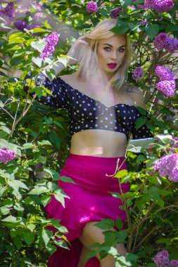 Çiçekli leylak çalıları arasında akıllı telefon kullanan sarışın bir kadının göz kamaştırıcı portresi. Bahar yaşam tarzı ve sosyal medya kavramı doğal ışıkta.