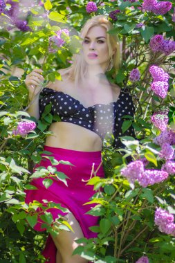 Yeşil bir bahçede mor leylak çiçeklerinin arasında poz veren sarışın bir kadın, üzerinde puantiyeli bir bluz ve parlak pembe etek, romantik bahar moda konsepti var..