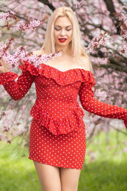 Kırmızı benekli elbisesiyle kiraz ağaçlarının altında poz veren sarışın kadın. Pembe çiçekli bahar moda portresi.