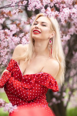 Kırmızı benekli elbiseli, bahar kiraz çiçekleriyle gülümseyen sarışın kadının güzel portresini kapat. Açık havada göz kamaştırıcı bir fotoğraf..