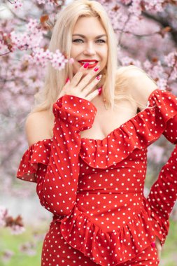 Kiraz çiçekleri arasında kırmızı benekli elbisesi olan, parlak manikürlü sarışın kadının oyuncu yakın çekimi. Bahar güzelliği ve moda portresi.