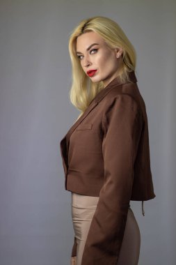 Omzunun üstünden bakan sarışın bir kadının profili, kahverengi ceket, kırmızı ruj, temiz gri stüdyo arka planı..