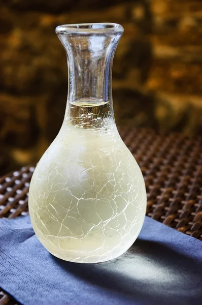 Ornate pitcher with cold white wine on a paper napkin over the wicker table in front of rough stone wall in rustic restaurant. Reflection. A game of light and shadow. Selective focus on the pitcher. — Stock Photo, Image