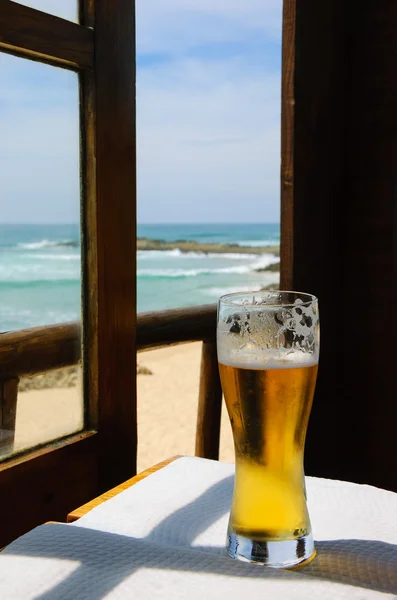 Koud biertje op café terras met uitzicht op het oceaan strand door het geopende raam. Algarve, Portugal. Een spel van licht een schaduw. — Stockfoto