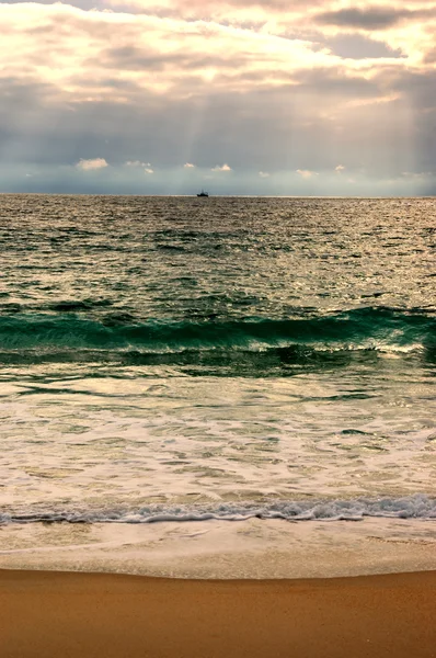 Dramática puesta de sol nublado con rayos de sol sobre el Océano Atlántico. Barco pesquero (rodeado de bandadas de gaviotas) navegando en el horizonte bajo los rayos del sol que brillan a través de las nubes. Nazare, Portugal . — Foto de Stock