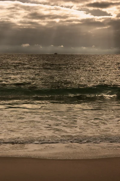 Dramática puesta de sol nublado con rayos de sol sobre la costa del Océano Atlántico. Barco pesquero navegando en el horizonte bajo los rayos del sol brillando a través de las nubes. Nazare, Portugal. Foto tonificada . — Foto de Stock