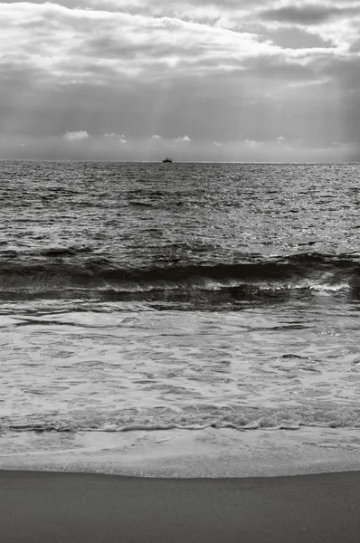 Dramatické zataženo západ slunce s paprsky přes Atlantský oceán. Rybářská loď (obklopen racků) plachtění na obzoru v slunečních paprsků zářící skrz mraky. Nazare, Portugalsko. Černá a bílá. — Stock fotografie