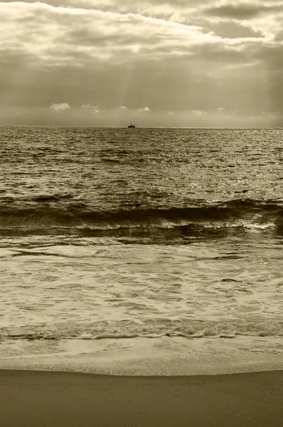 Dramatiska grumlig solnedgången med solstrålar över Atlanten. Fiske fartyg (omgiven av flock måsar) segling på horisonten i solstrålar glödande genom molnen. Nazaré, Portugal. Sepia foto. — Stockfoto