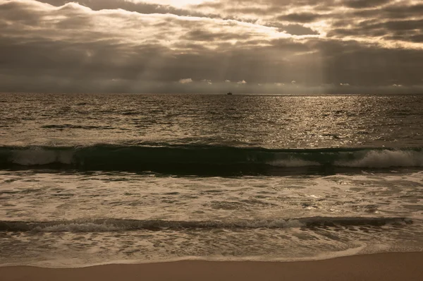 Dramática puesta de sol nublado con rayos de sol sobre la costa del Océano Atlántico. Barco pesquero navegando en el horizonte bajo los rayos del sol brillando a través de las nubes. Nazare, Portugal. Foto tonificada . — Foto de Stock