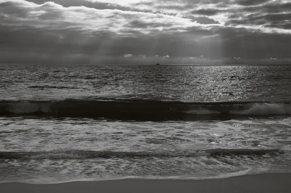 Dramatické zataženo západ slunce s paprsky na pobřeží Atlantského oceánu. Rybářská loď plující na obzoru pod paprsky slunce svítí skrz mraky. Nazare, Portugalsko. Věku fotografie. Černá a bílá. — Stock fotografie