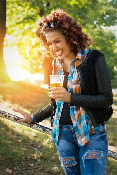 Glad ung kaukasiska rödhårig kvinna i parken på solig dag håller ett glas öl — Stockfoto