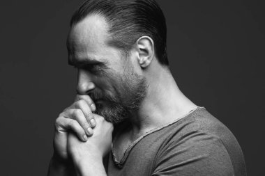 Hope concept. Profile portrait of 40-year-old man praying over dark gray background. Hair brushed back. Rocker, biker style. Hands near face. Close up. Monochrome studio shot