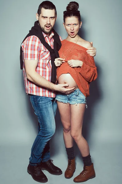 Stylish pregnancy & family concept: portrait of disturbed couple of hipsters (husband and wife) in trendy casual clothing, eyewear posing over gray background. Urban street style. Studio shot — Stock Photo, Image