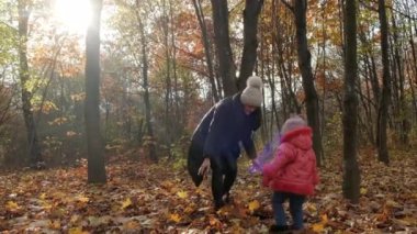 Genç bir kadın ve küçük kızı şehir parkına sarı sonbahar yaprakları fırlatıyorlar. Yavaş çekim.