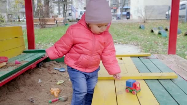 Leksaksflickan leker med leksaksbilen i sandlådan. Långsamma rörelser. — Stockvideo