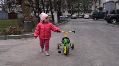Küçük kız cadde boyunca üç tekerlekli bisikletin yanında yürüyor. Yavaş çekim.