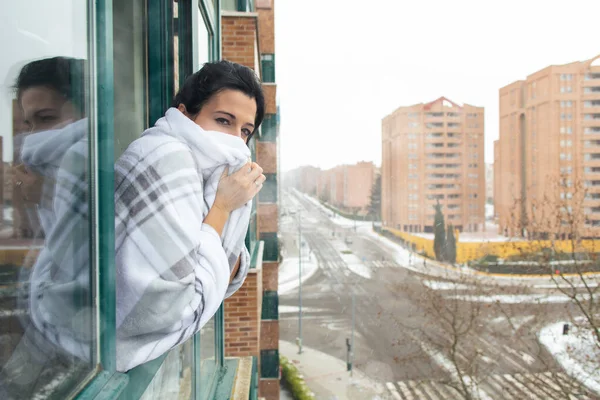 Woman wrapped in a warm white blanket looking out the window during a cold winter snow storm.