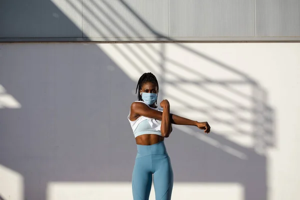 外コロナウイルスパンデミックの下で顔マスクトレーニングを持つ女性都市部の選手を動機付け 肩は走る前に暖かい運動を伸ばす — ストック写真